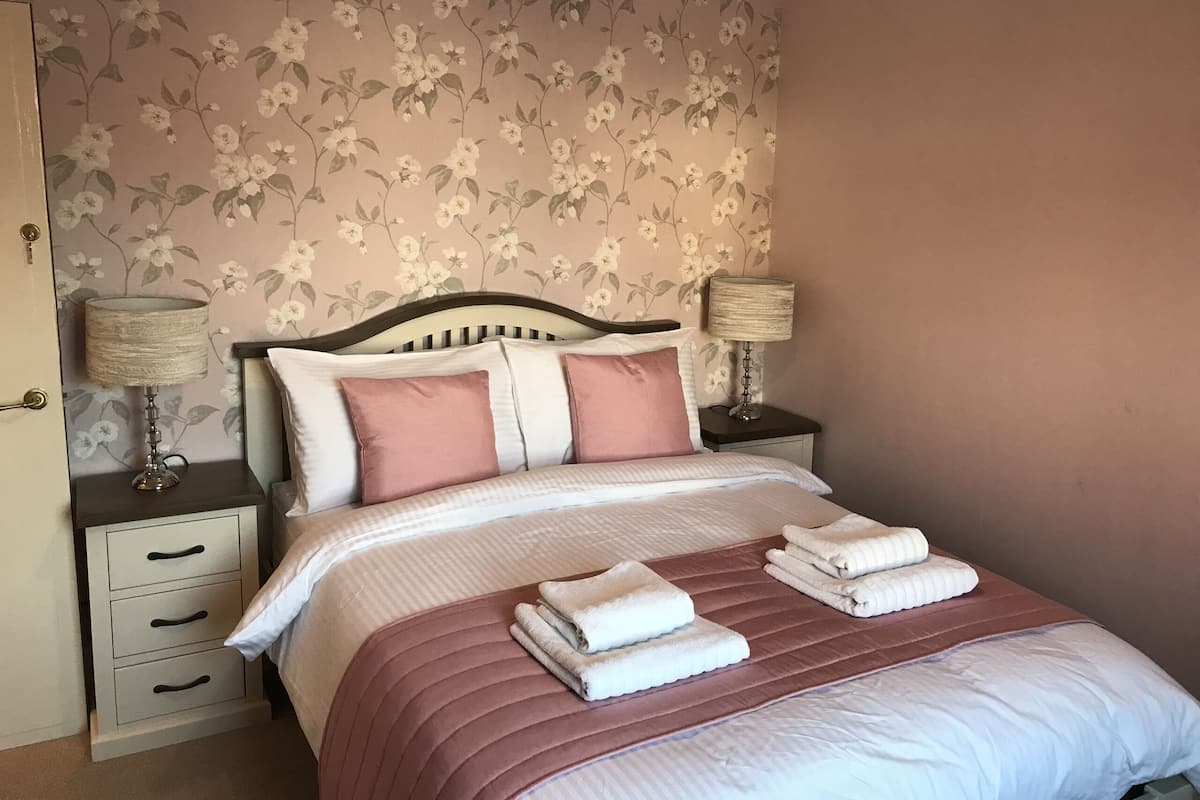 Cama bem arrumada com roupa de cama rosa e branca, duas luminárias de cabeceira e um papel de parede floral.