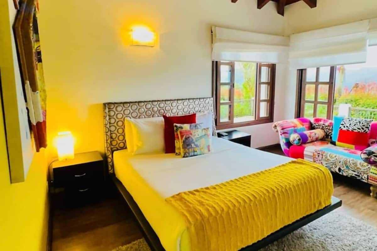 Une chambre avec un plafond en bois, un lit recouvert d’une couverture jaune, une table de chevet et une fenêtre donnant sur un paysage verdoyant.
