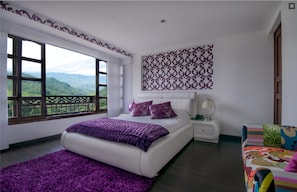 Un dormitorio con un ventanal grande, una cama con ropa de cama morada, un muro estampado y un sofá colorido.