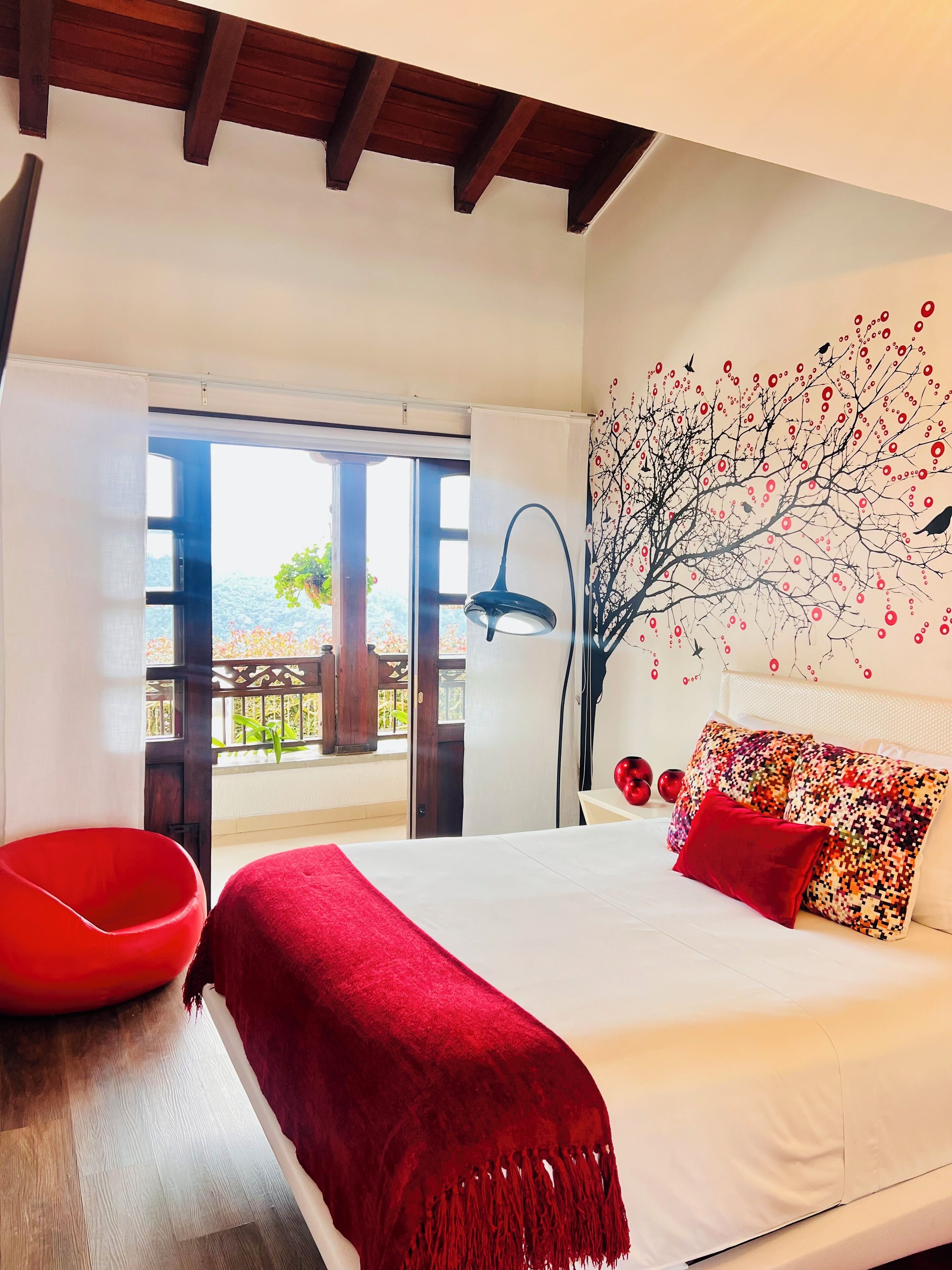 A bedroom with a bed, a red chair, a balcony with a view, and a wall mural.