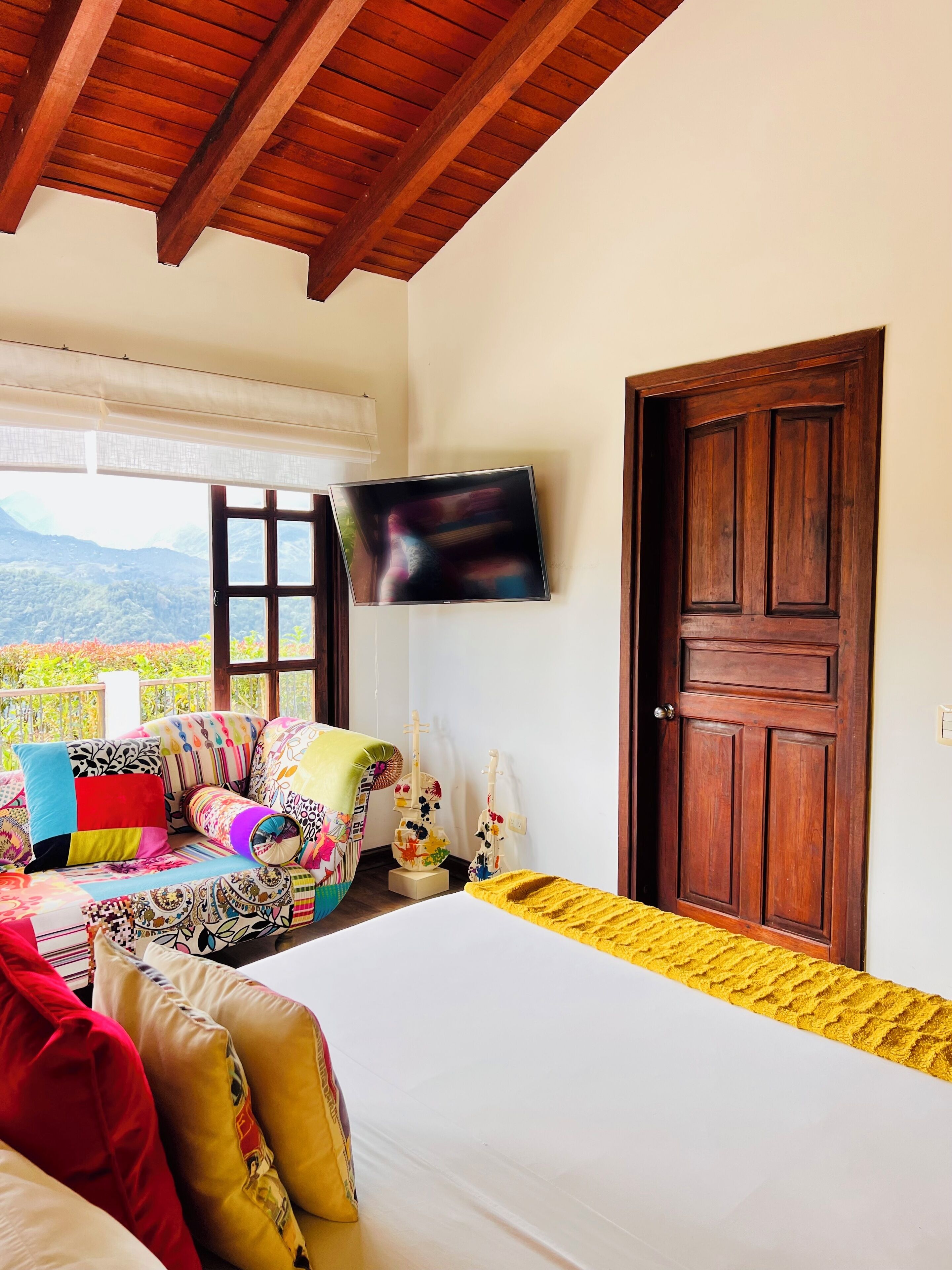 Un dormitorio con cama, almohadones coloridos, un sofá, una puerta de madera y un televisor en la pared.