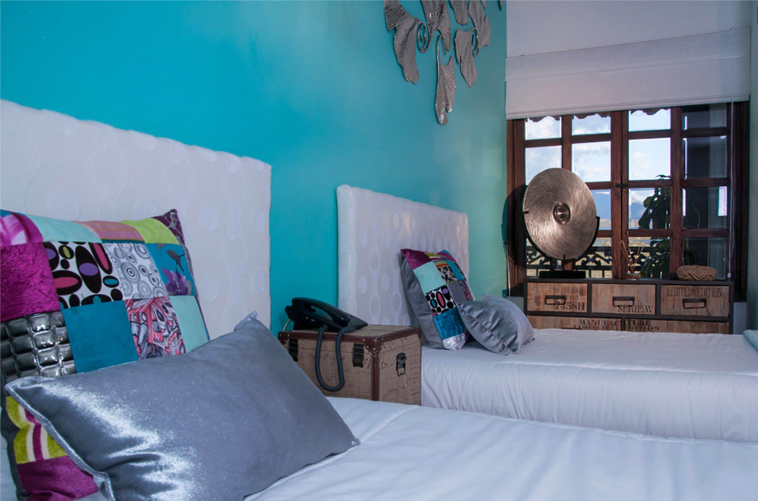 Una habitación con dos camas, almohadas coloridas, una maleta y una ventana con vistas.