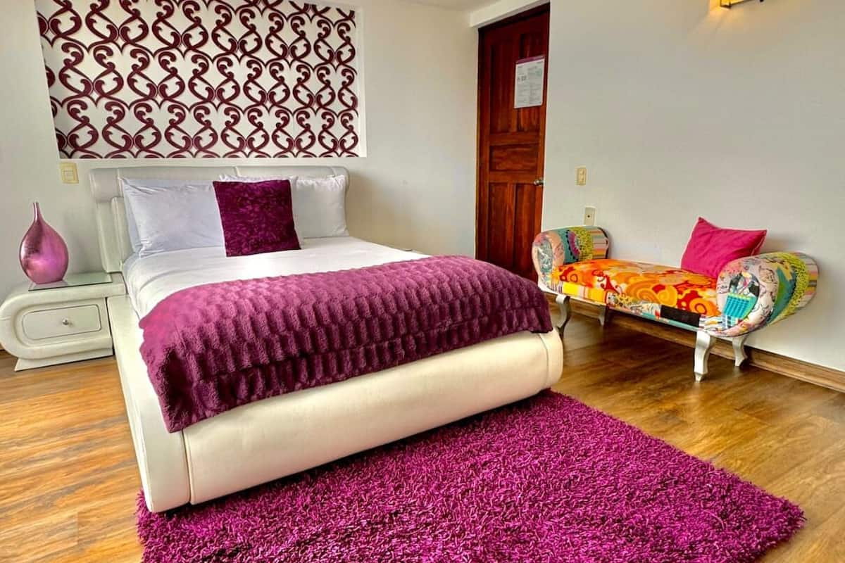 Une chambre avec un grand lit, une table de chevet, un panneau décoratif au mur, un canapé agrémenté de coussins colorés et un plancher en bois.
