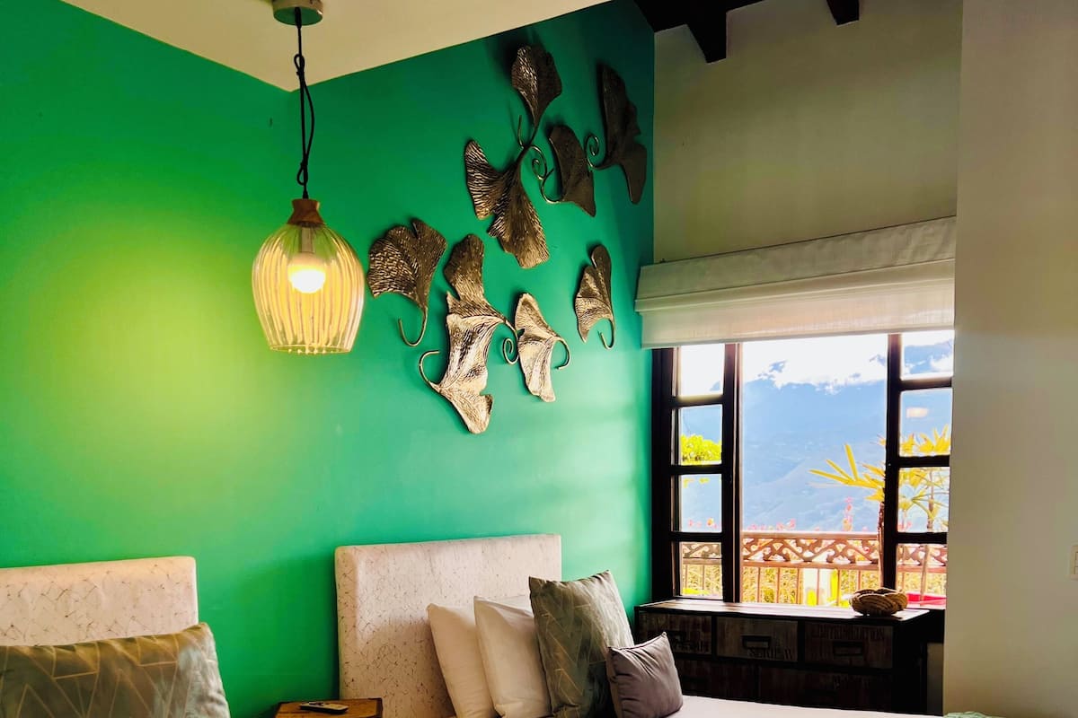 Une chambre avec deux lits, un mur vert, des éléments décoratifs muraux, un luminaire suspendu et une fenêtre donnant sur les montagnes.