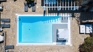 An aerial view of a swimming pool with a stone wall surrounding it, a covered seating area with cushions, and a small garden with flowers. - The One Mykonos (Mykonos)