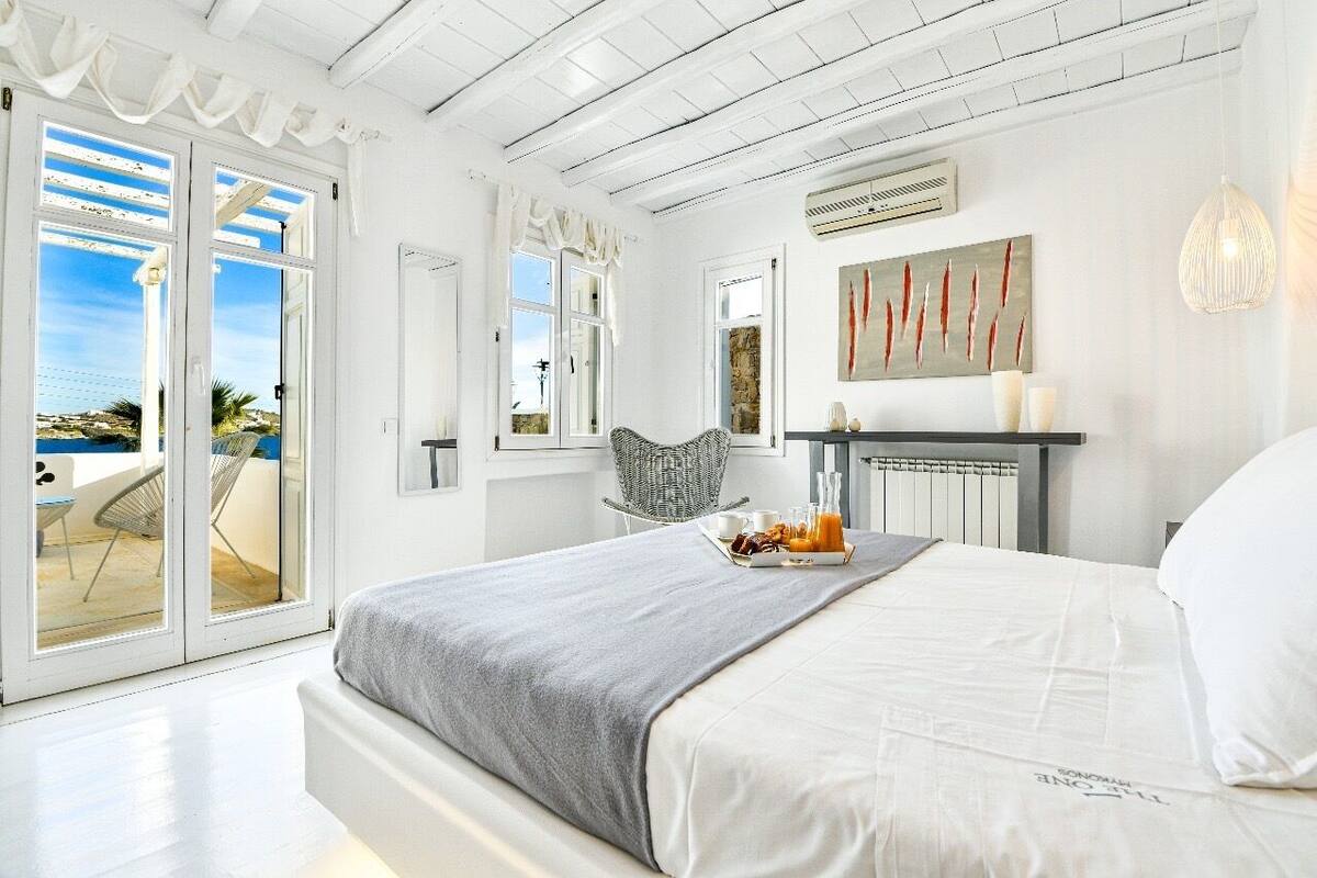 A bedroom with a bed, a breakfast tray, a wicker chair, and a view of the sea through large sliding doors.