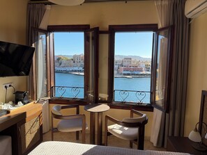 Une chambre d’hôtel comprenant un lit, un bureau avec un ordinateur, deux chaises, une petite table et un téléviseur. De grandes fenêtres offrent une vue sur le bord de l’eau, avec des bâtiments et un pont.