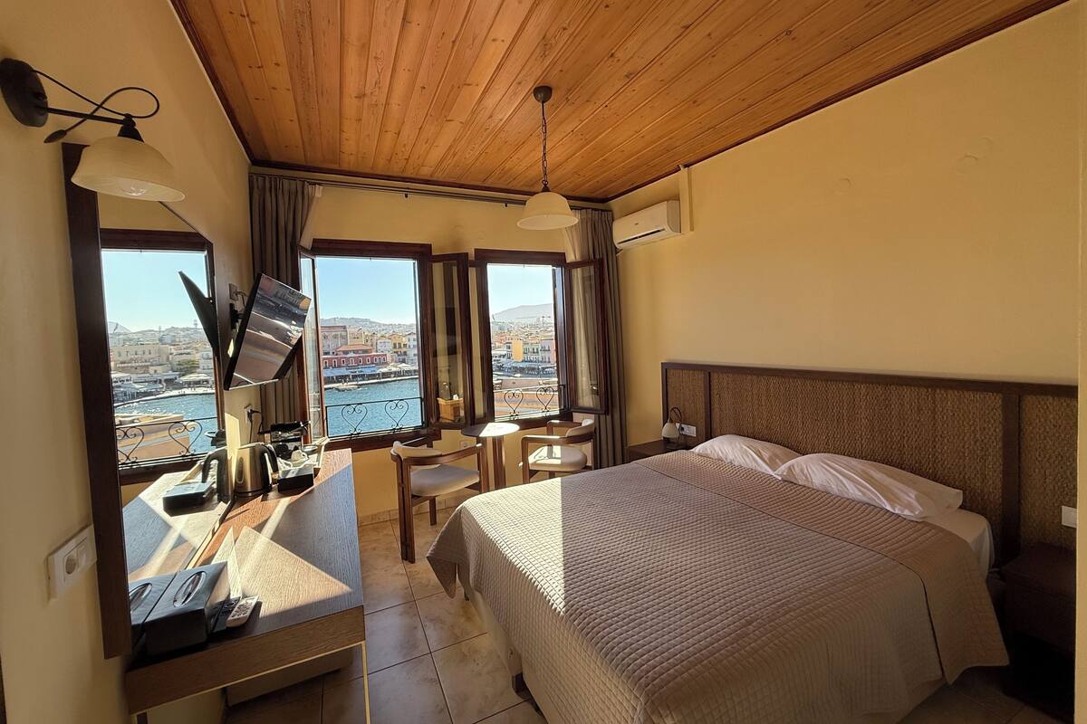 A hotel room with a bed, desk, and large windows offering a view of a cityscape and water.