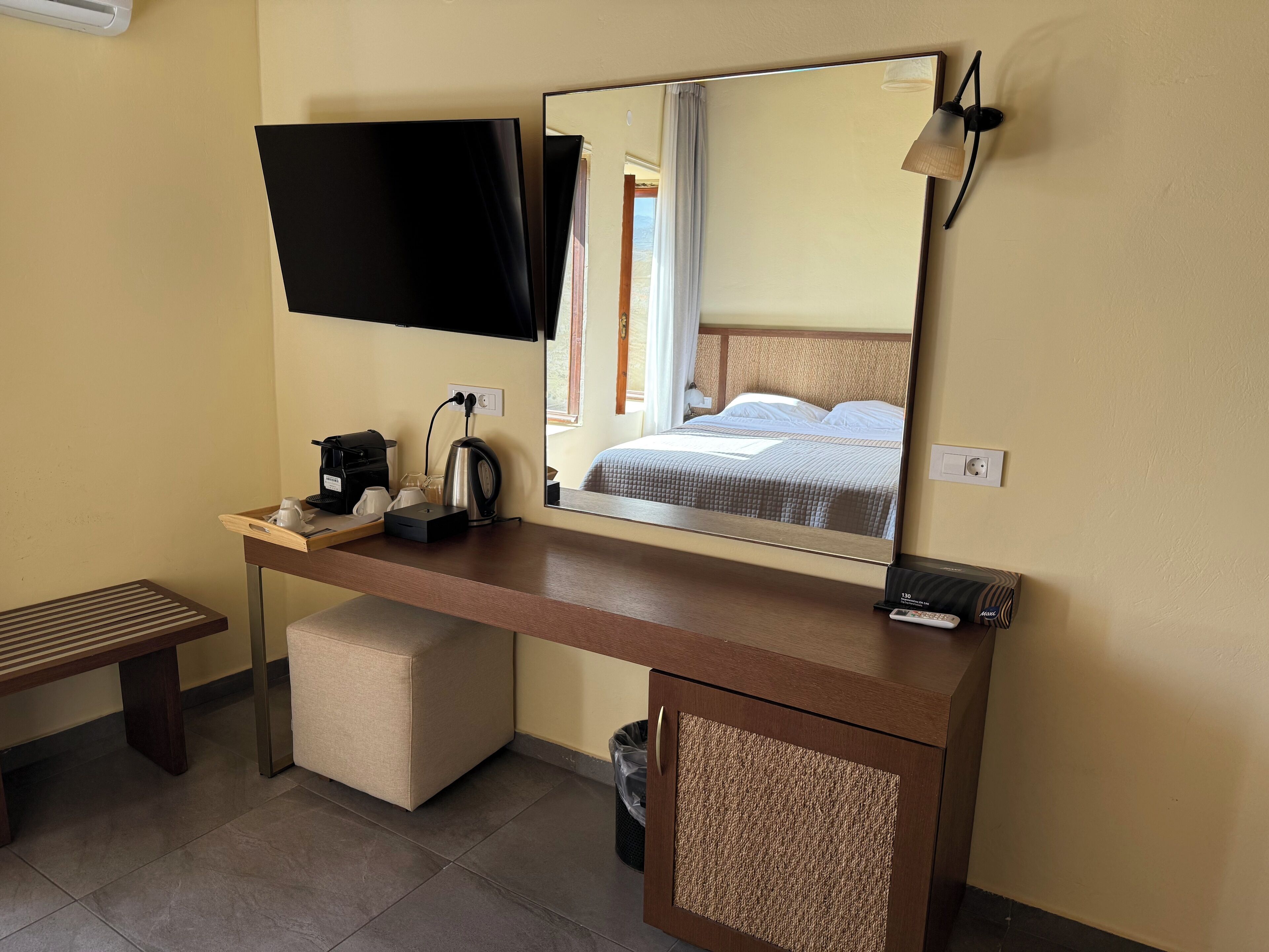 A hotel room with a flat-screen TV mounted on the wall, a wooden desk with a coffee maker, a trash bin, and a bench.