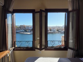 Ένα δωμάτιο με ένα κρεβάτι και δύο παράθυρα που έχουν θέα σε ένα λιμάνι, με κτίρια και μια προβλήτα.