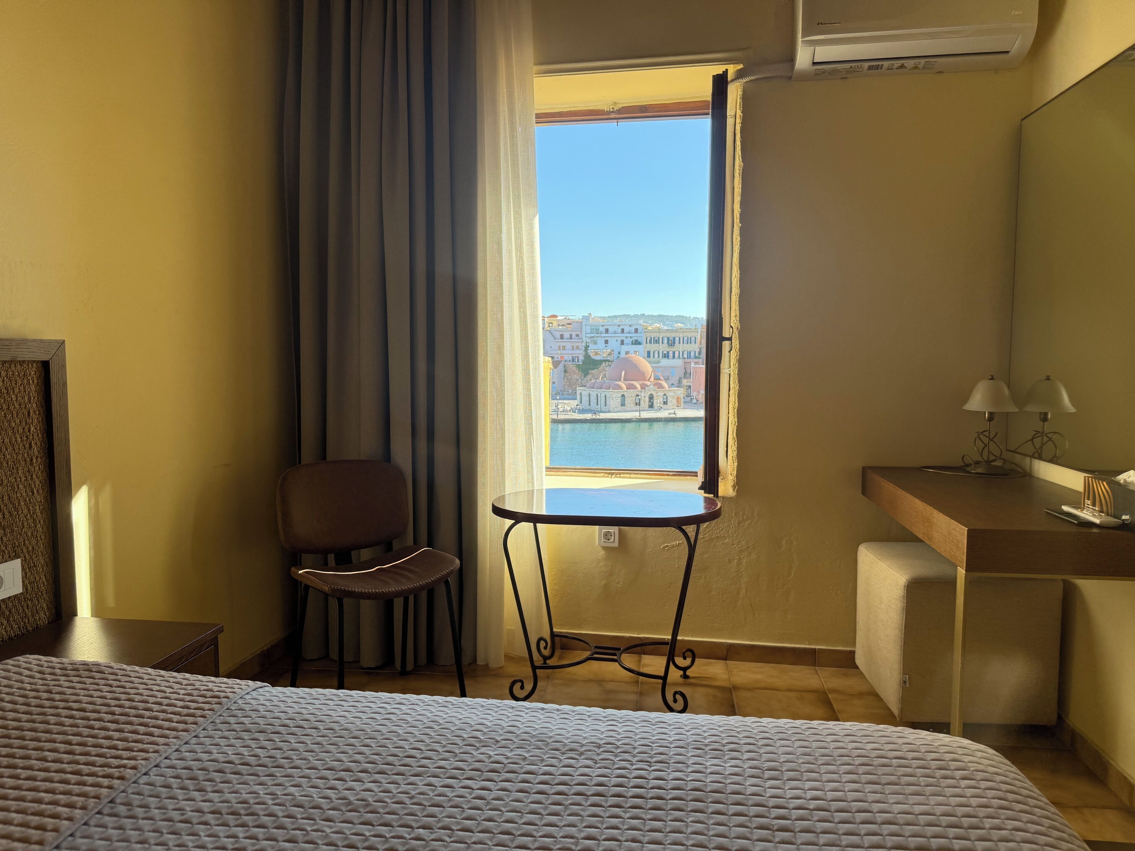A hotel room with a bed, a chair, a small table, a desk, and a window with a view of buildings and water.