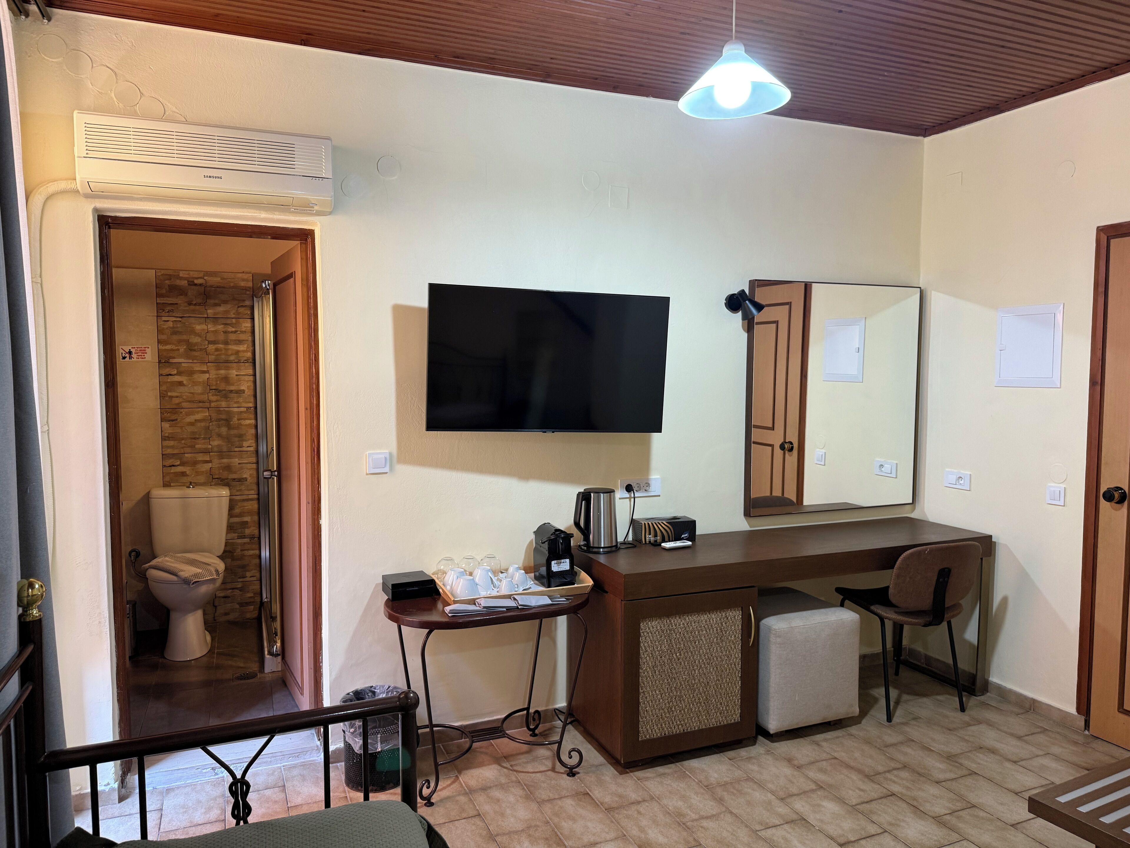A hotel room with a flat-screen TV, a desk with a chair, a small refrigerator, a coffee maker, and a trash bin.