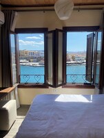 Ένα δωμάτιο με ένα κρεβάτι, ένα παράθυρο με θέα σε ένα λιμάνι και κτίρια, και έναν ανεμιστήρα οροφής.