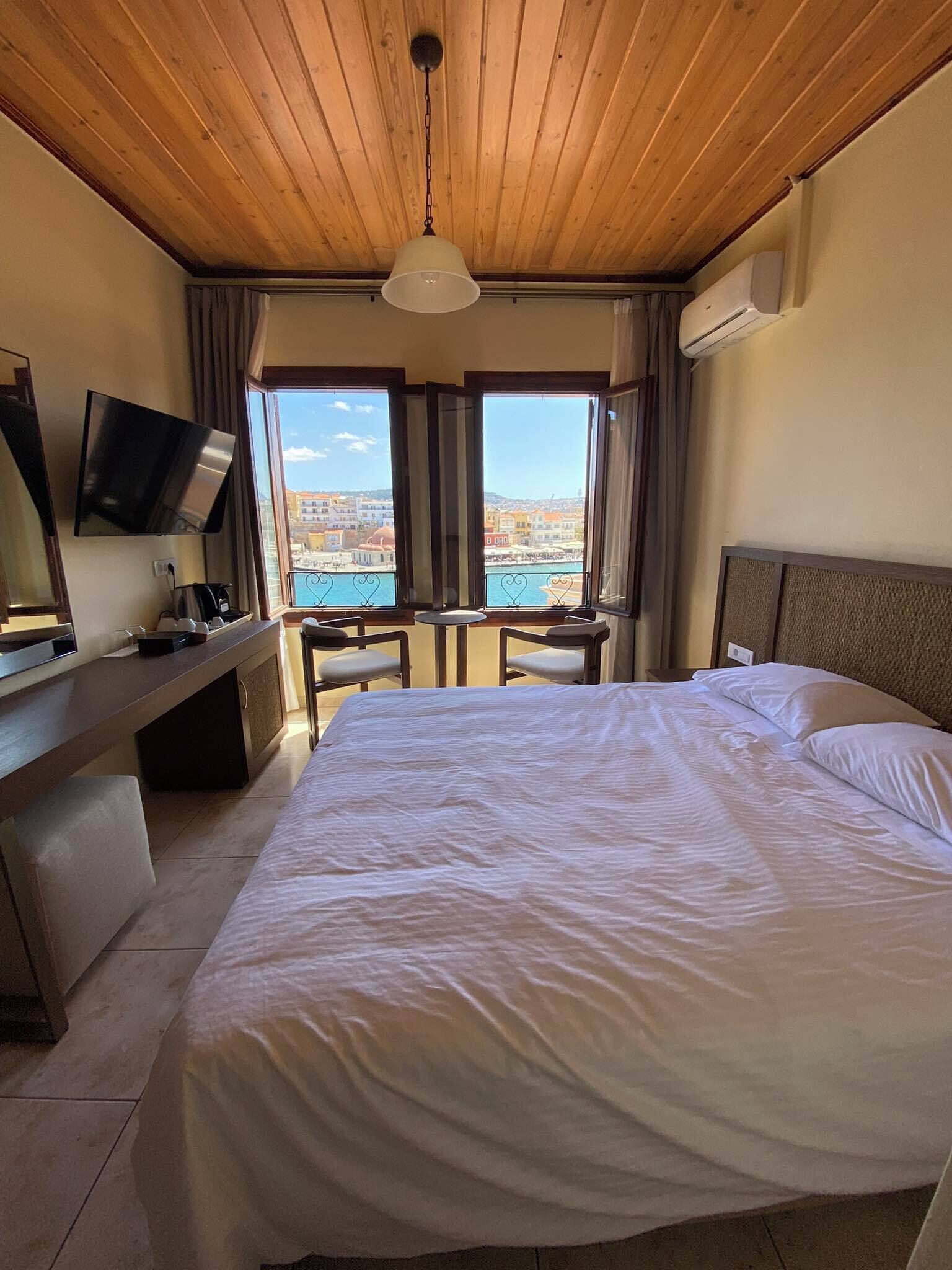 A hotel room with a large bed, a desk with a chair, a TV, and a view of the sea.