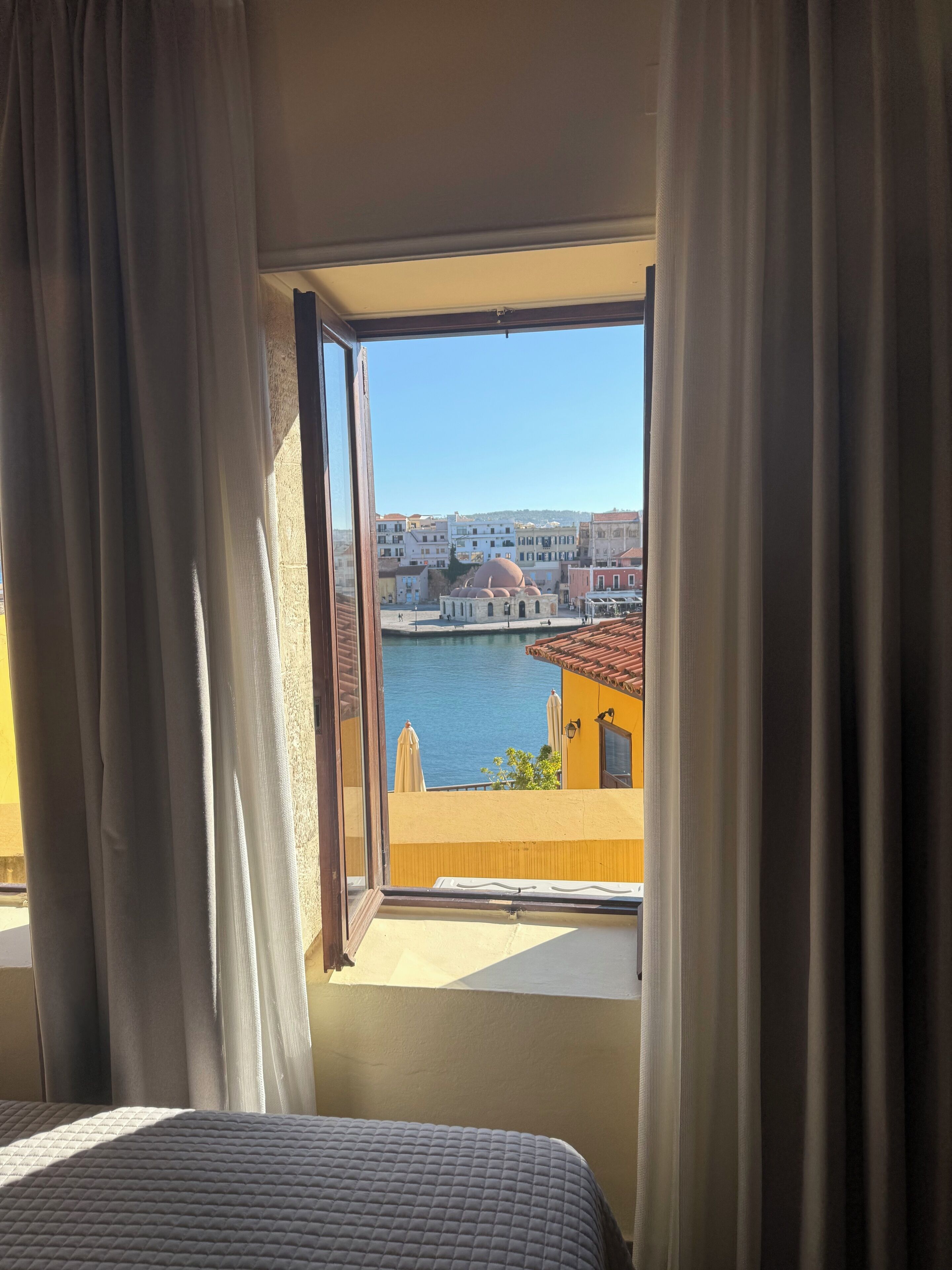 A room with a bed, a window with curtains, and a view of a cityscape with buildings and a body of water.