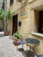 Μια αυλή με μια πινακίδα που γράφει «ΞΕΝΟΔΟΧΕΙΟ ΕΛΕΝΑ», γλάστρες με φυτά, ένα μεταλλικό τραπέζι και μια καρέκλα, και ένα δάπεδο από τούβλα.