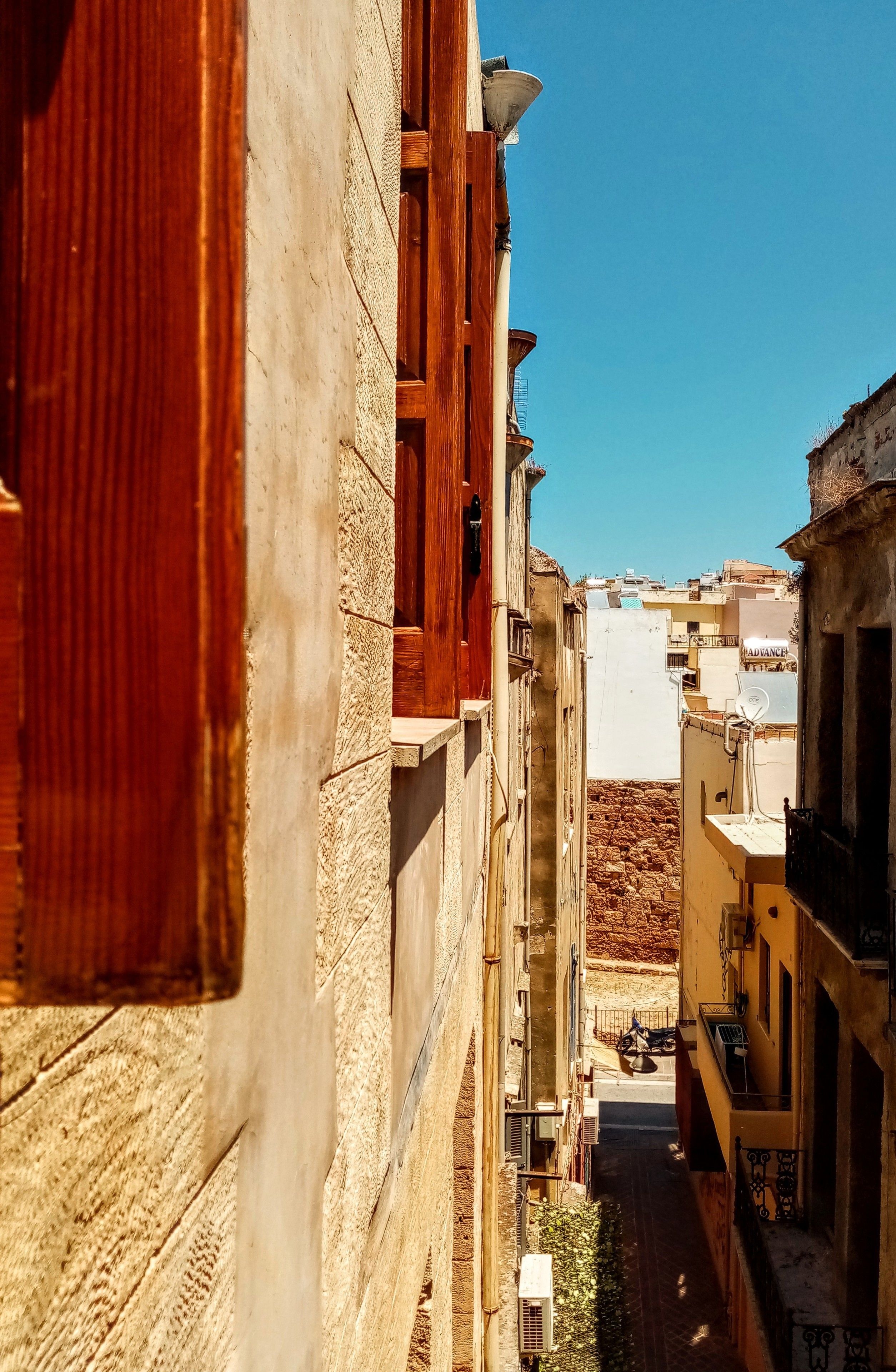 A narrow street with buildings on either side, a clear blue sky, and a distant view of a hill.