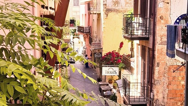 Une rue étroite, bordée de bâtiments des deux côtés, avec des balcons ornés de plantes et des espaces aménagés pour s’asseoir à l’extérieur.