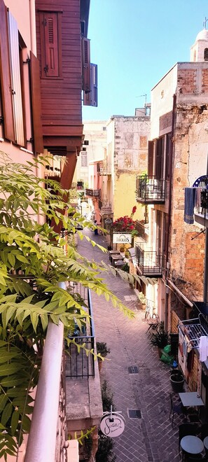 A narrow street with buildings on either side, featuring balconies with plants and outdoor seating. - Palazzo di Pietro (Chania)