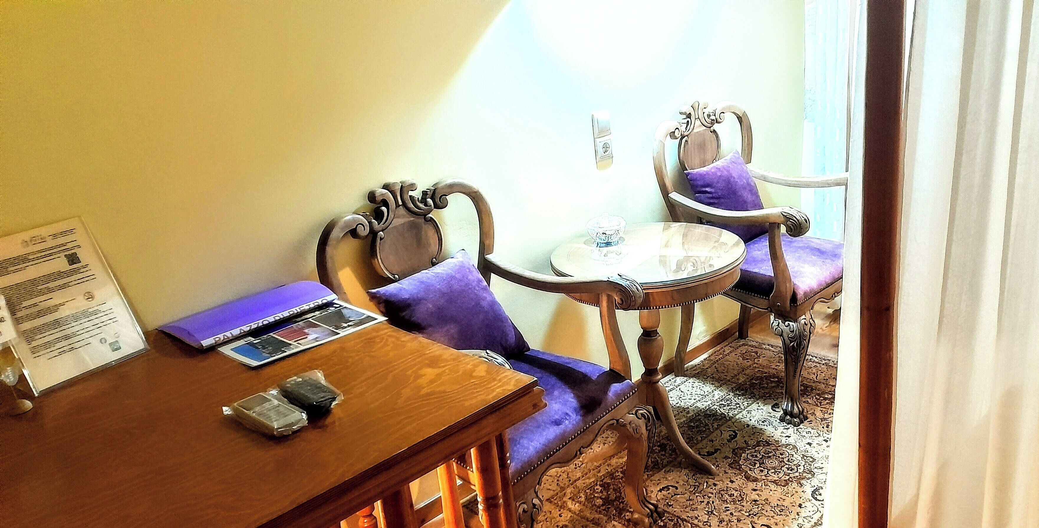 A room with a wooden table and two chairs, a small round table with a glass top, and a purple cushion on one of the chairs.