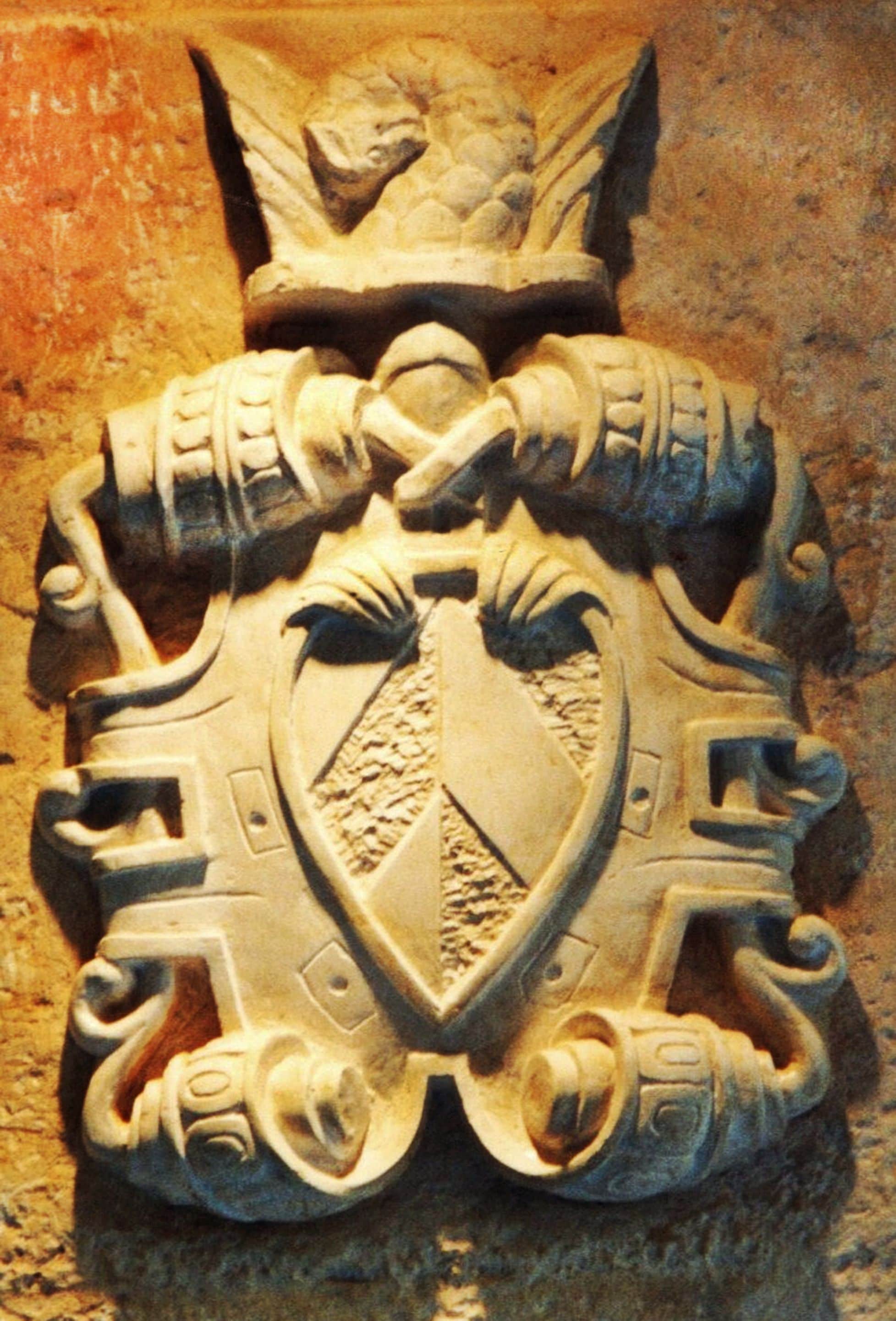 Een stenen embleem met ingewikkelde gravures, waaronder een centraal schild en omliggende decoratieve details.