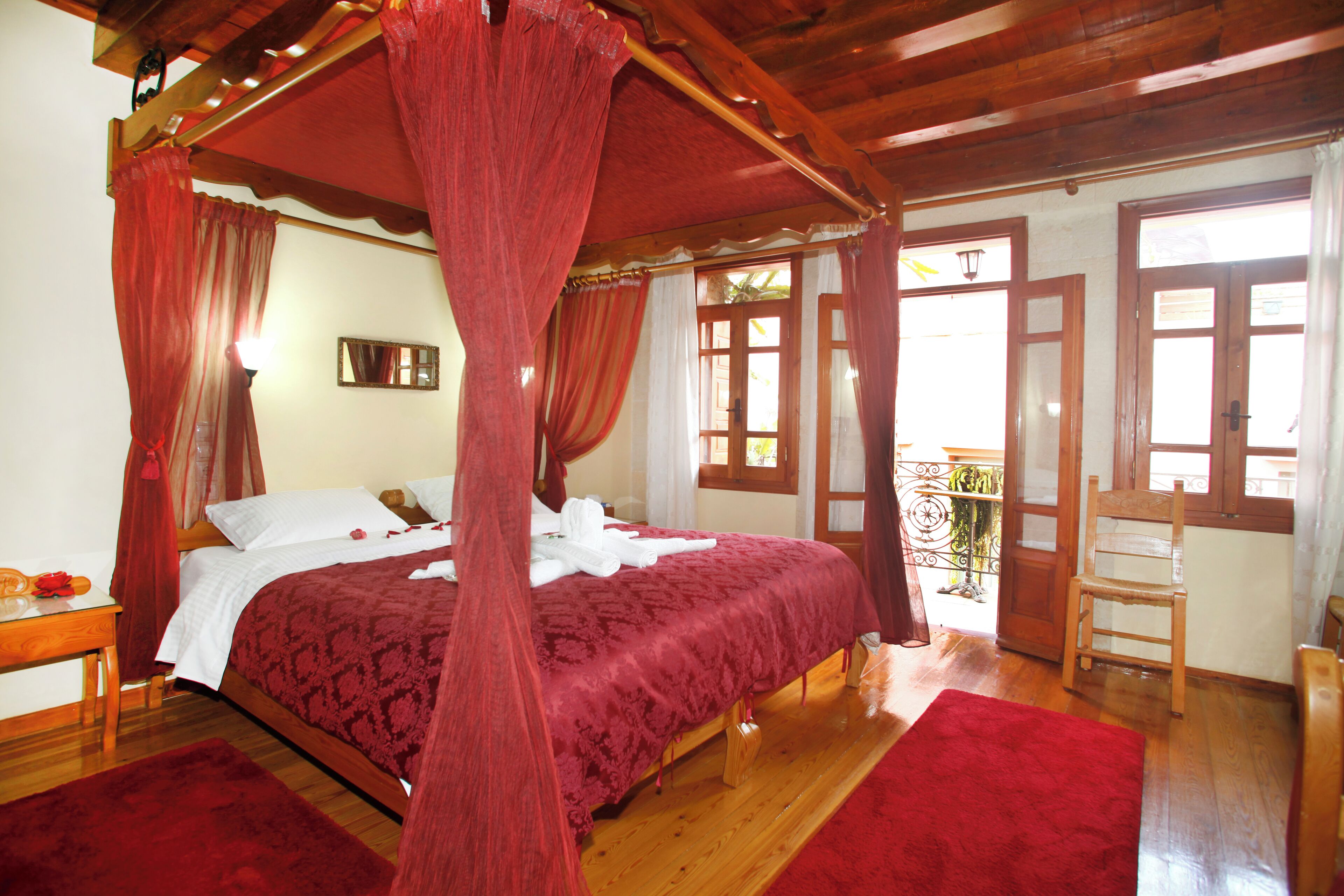 A four-poster canopy bed with a red bedspread, wooden flooring, and a balcony with a wooden chair.