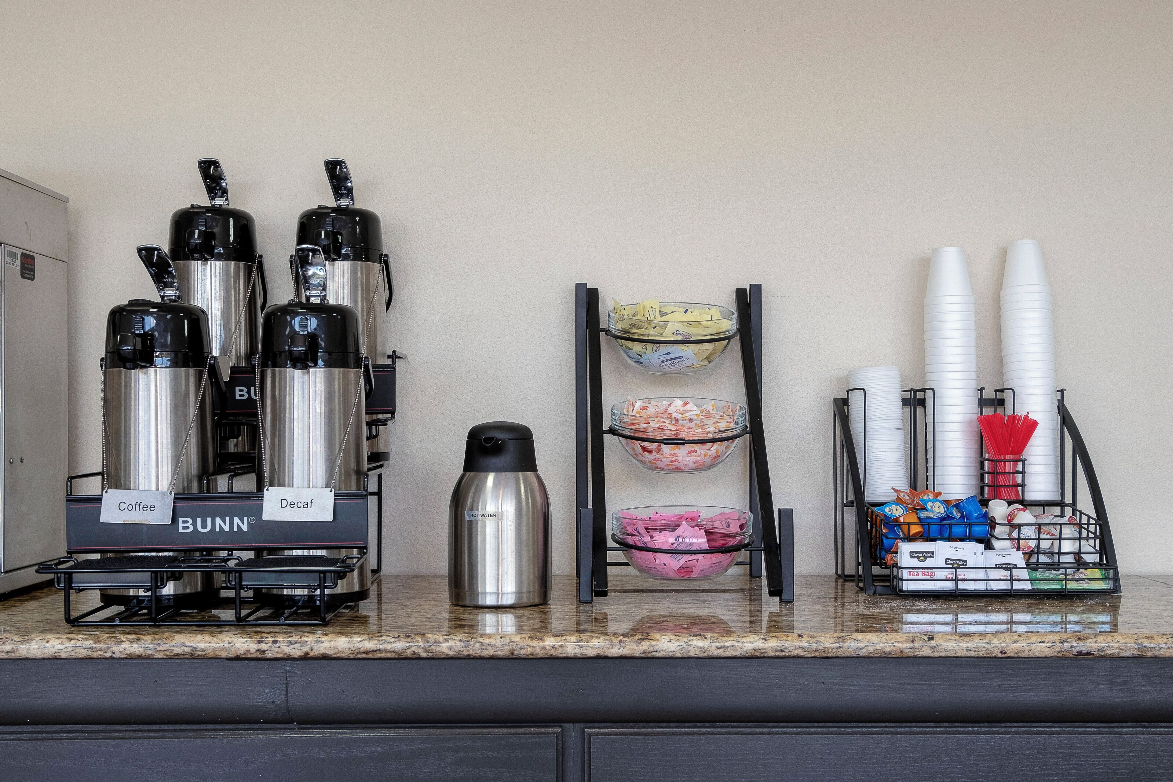 A kitchenette with coffee and tea dispensers, a water bottle, and a rack holding cups and utensils.