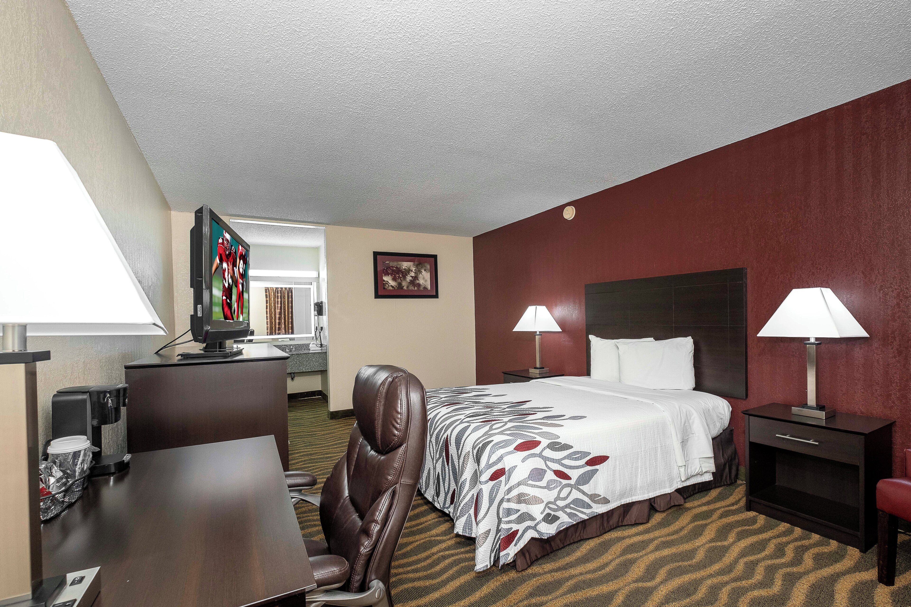 Ein Hotelzimmer mit einem Bett, zwei Nachttischlampen, einem Schreibtisch mit einem Computer, einem Sessel und einem Fernseher auf einem Tisch.