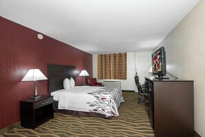 A hotel room with a bed, two bedside lamps, a television, a desk with a computer, and a patterned carpet.