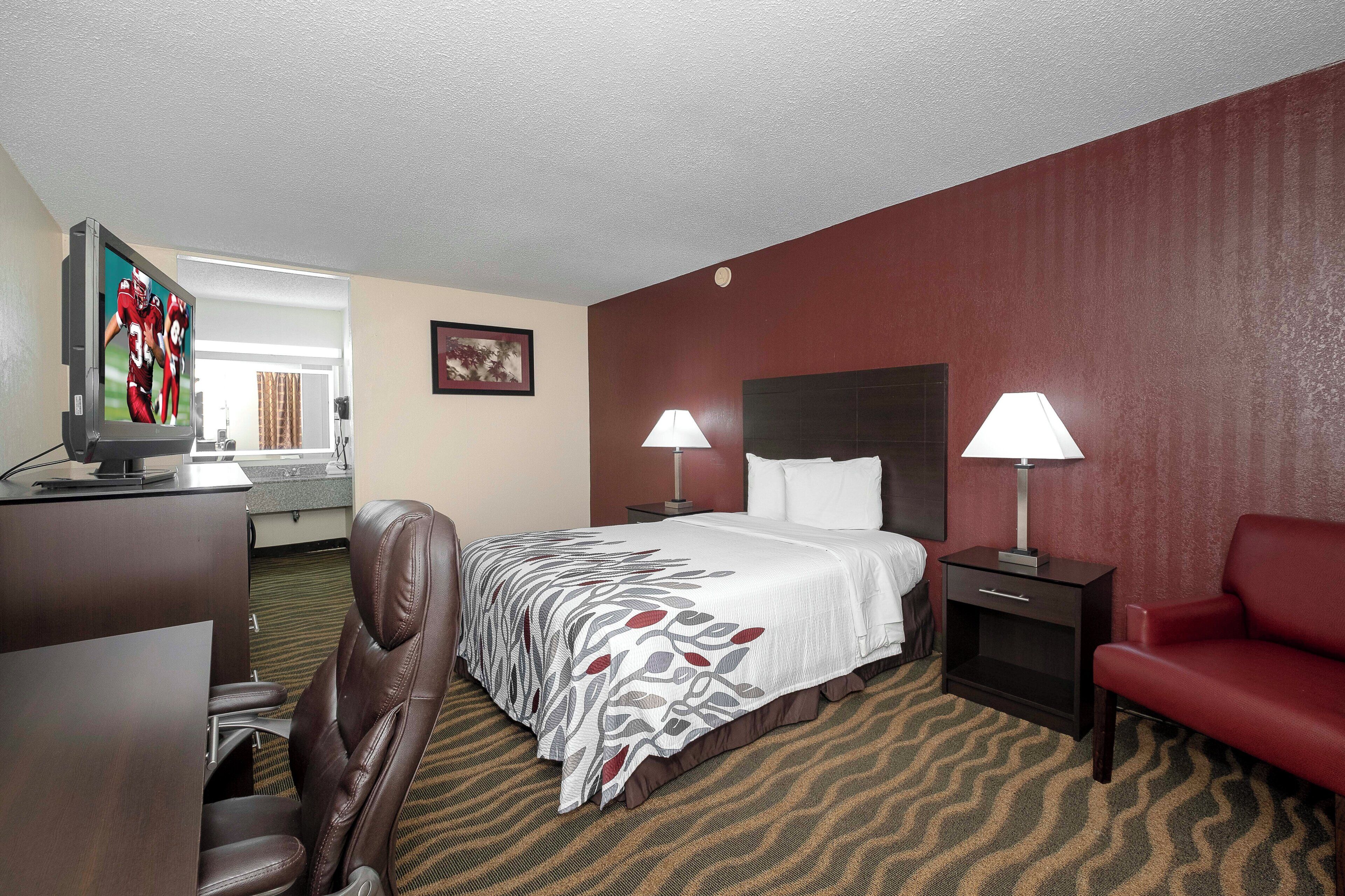 A hotel room with a bed, two bedside lamps, a desk with a TV, a chair, and a red couch.