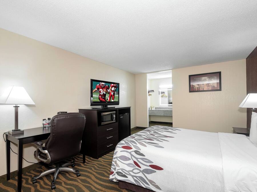 Quarto de hotel com cama, escrivaninha, cadeira, televisão e banheiro visível por uma porta aberta.