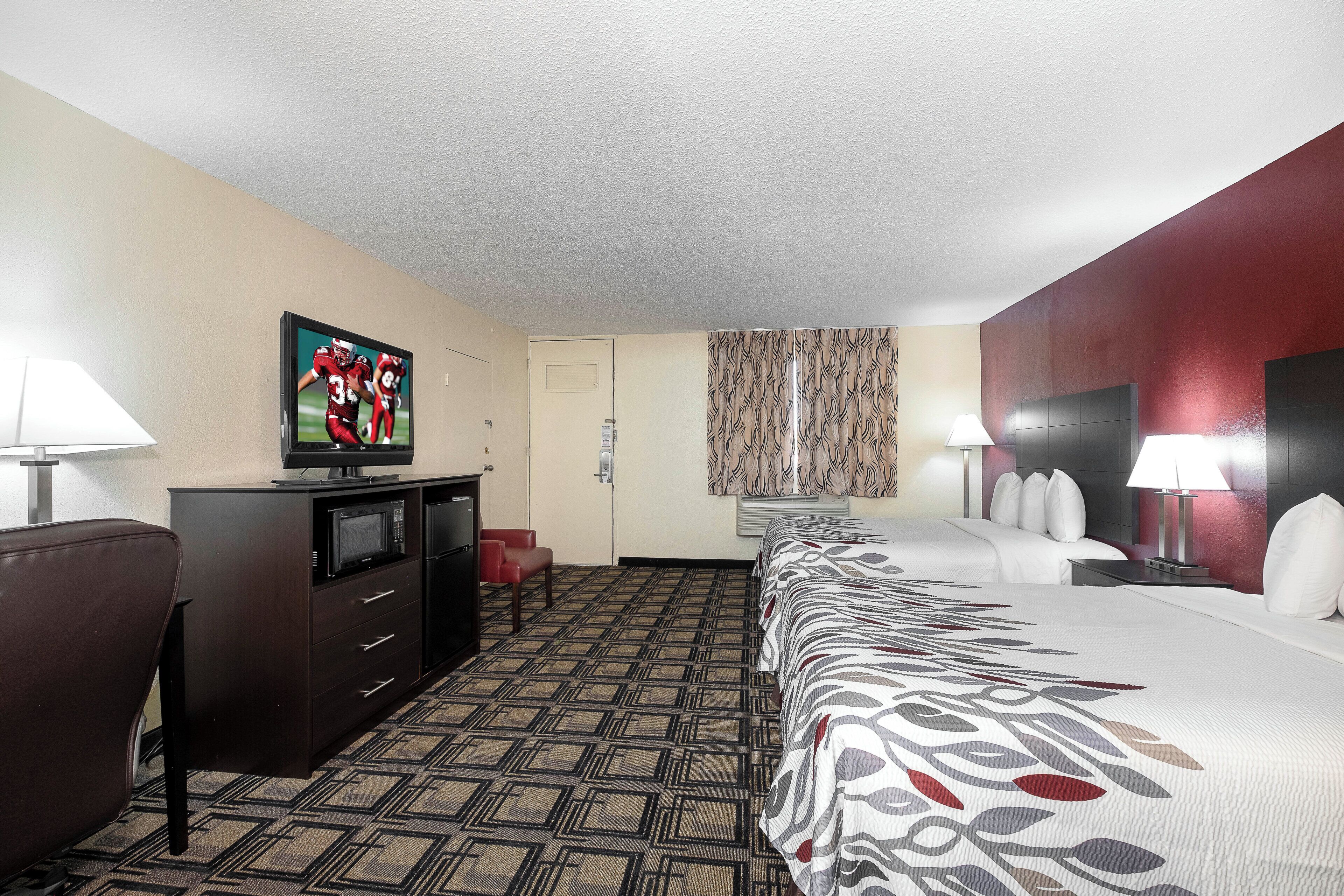A hotel room with a bed, a TV, a desk, a chair, a lamp, and a patterned carpet.