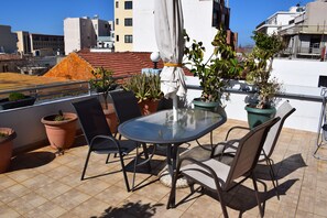 A rooftop terrace with a glass table and chairs, surrounded by potted plants and a white umbrella.