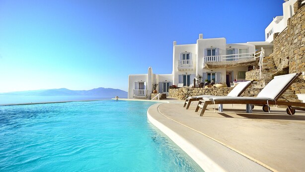 A poolside area with lounge chairs, a clear blue pool, and a white building with balconies overlooking the sea. - Villa Adella by Mermaid Luxury Villas (Mykonos)