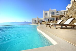 Ein Poolbereich mit Liegestühlen, ein klares, blaues Schwimmbecken und ein weißes Gebäude mit Balkonen mit Blick auf das Meer.