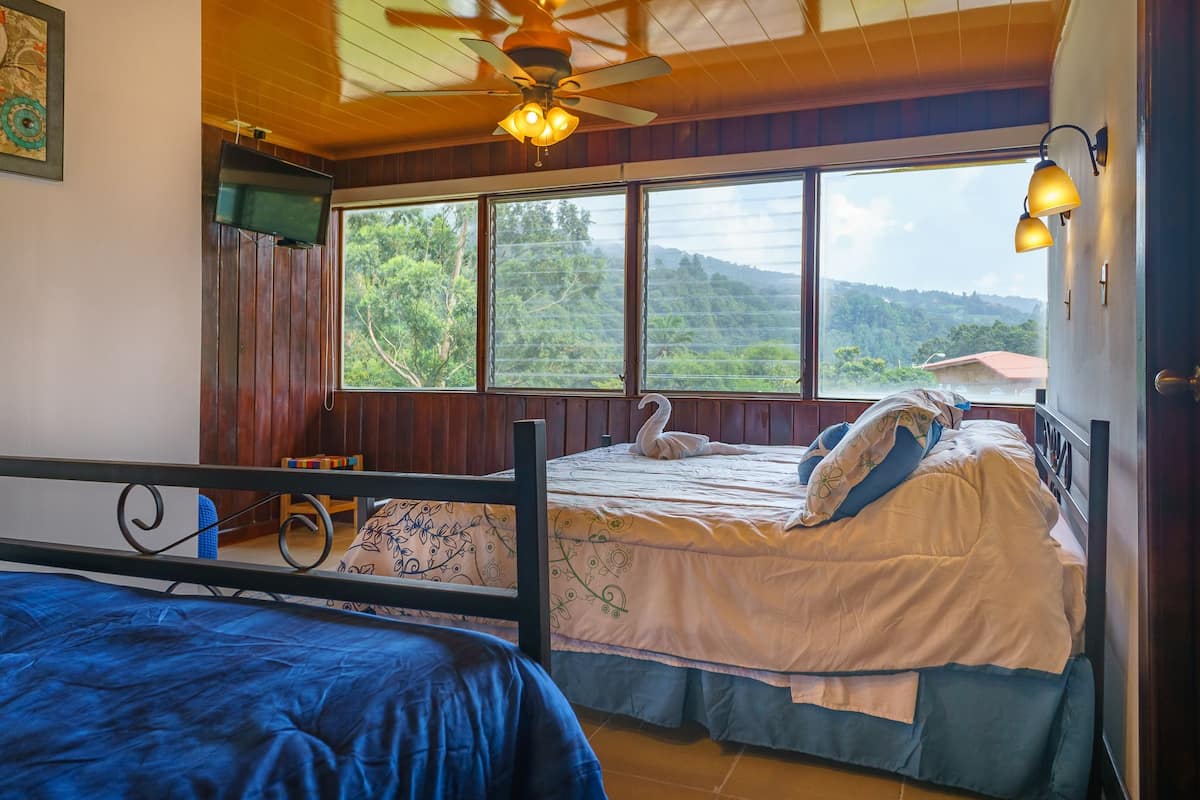 A bedroom with a bed, a chair, a ceiling fan, and a window with a view of greenery.