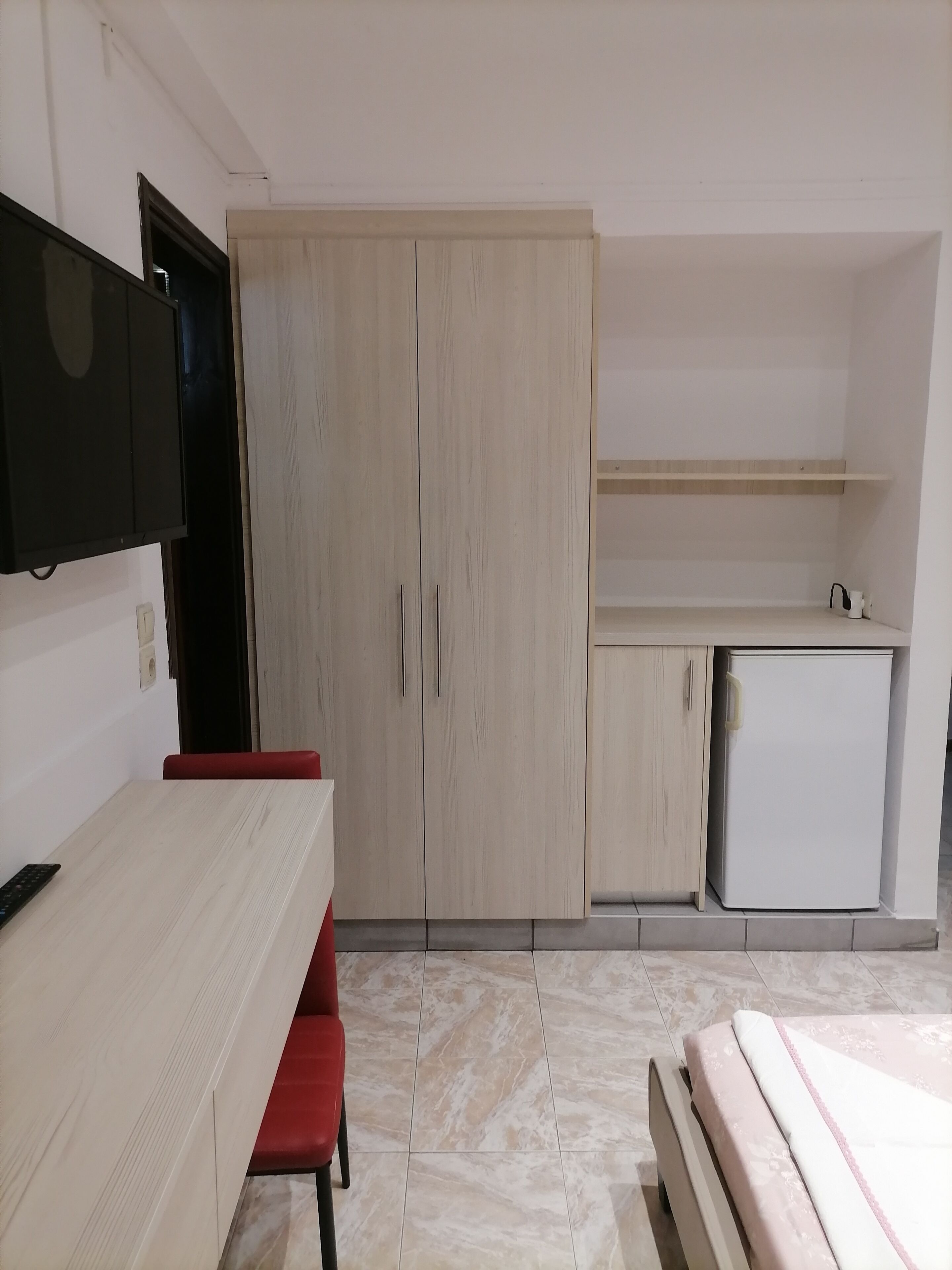 A compact room with a bed, a desk with a chair, a small kitchenette, and a wardrobe.