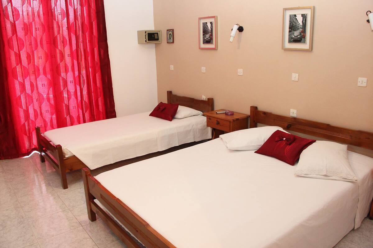 Two single beds with wooden frames, white linens, and red pillows. There are bedside tables with lamps and a small shelf with a picture frame.
