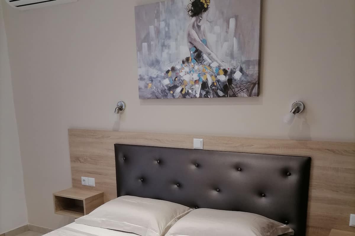 A bedroom with a bed, a headboard, a nightstand, an air conditioning unit, and a painting on the wall.