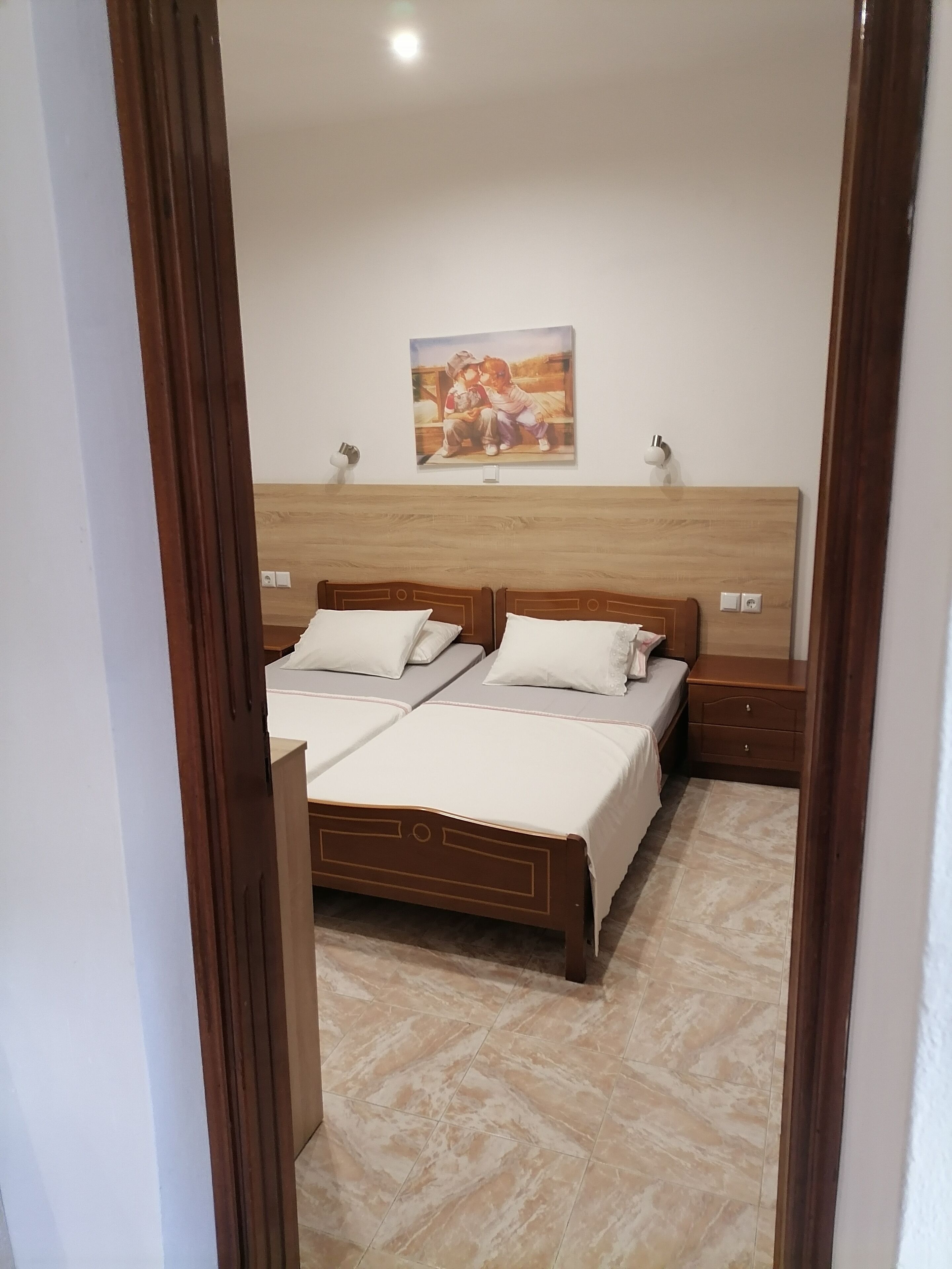 Une chambre avec deux lits, une tête de lit en bois, un tableau au mur et un sol carrelé.