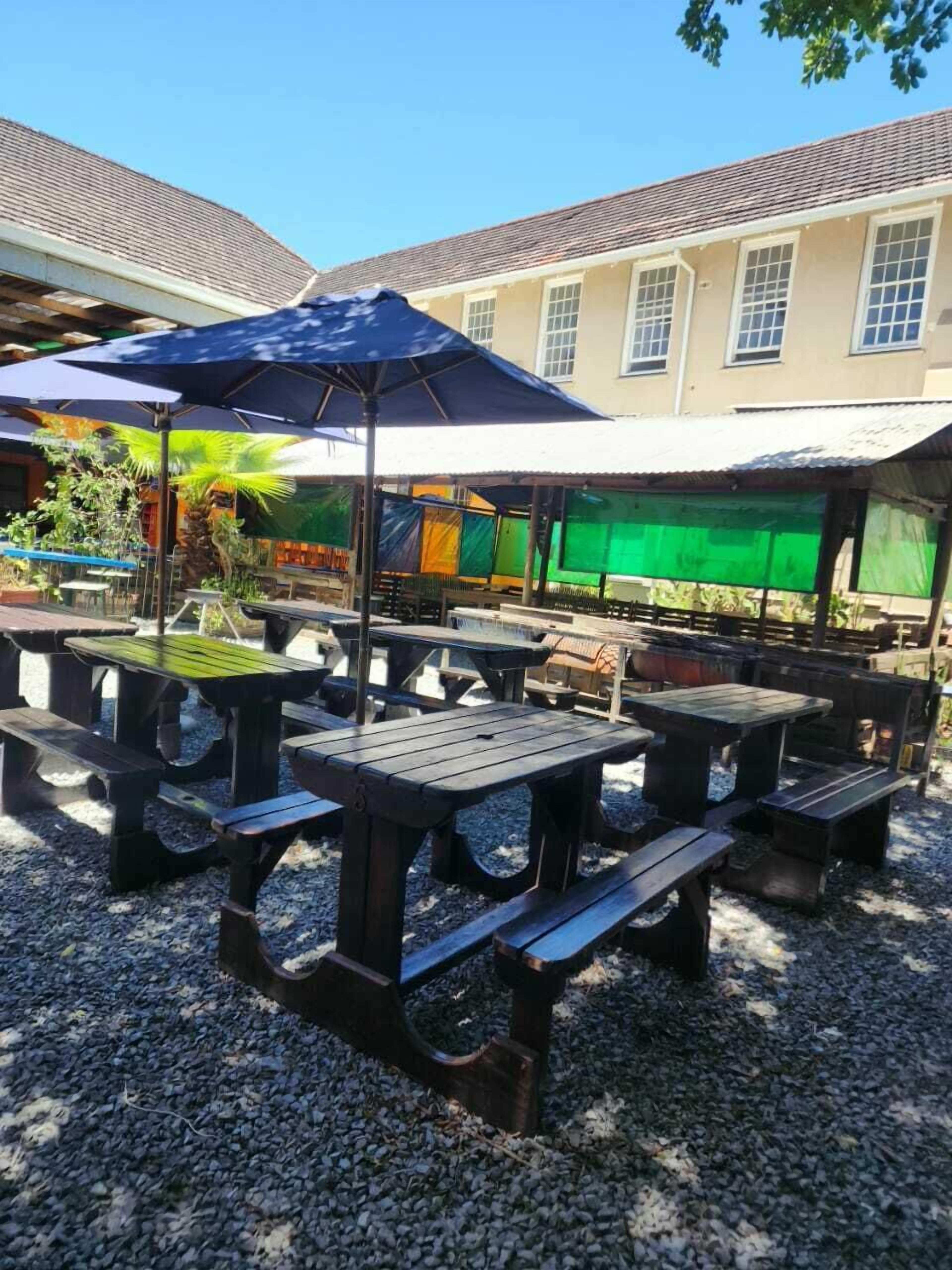 An outdoor seating area with picnic tables and umbrellas, surrounded by gravel and greenery.