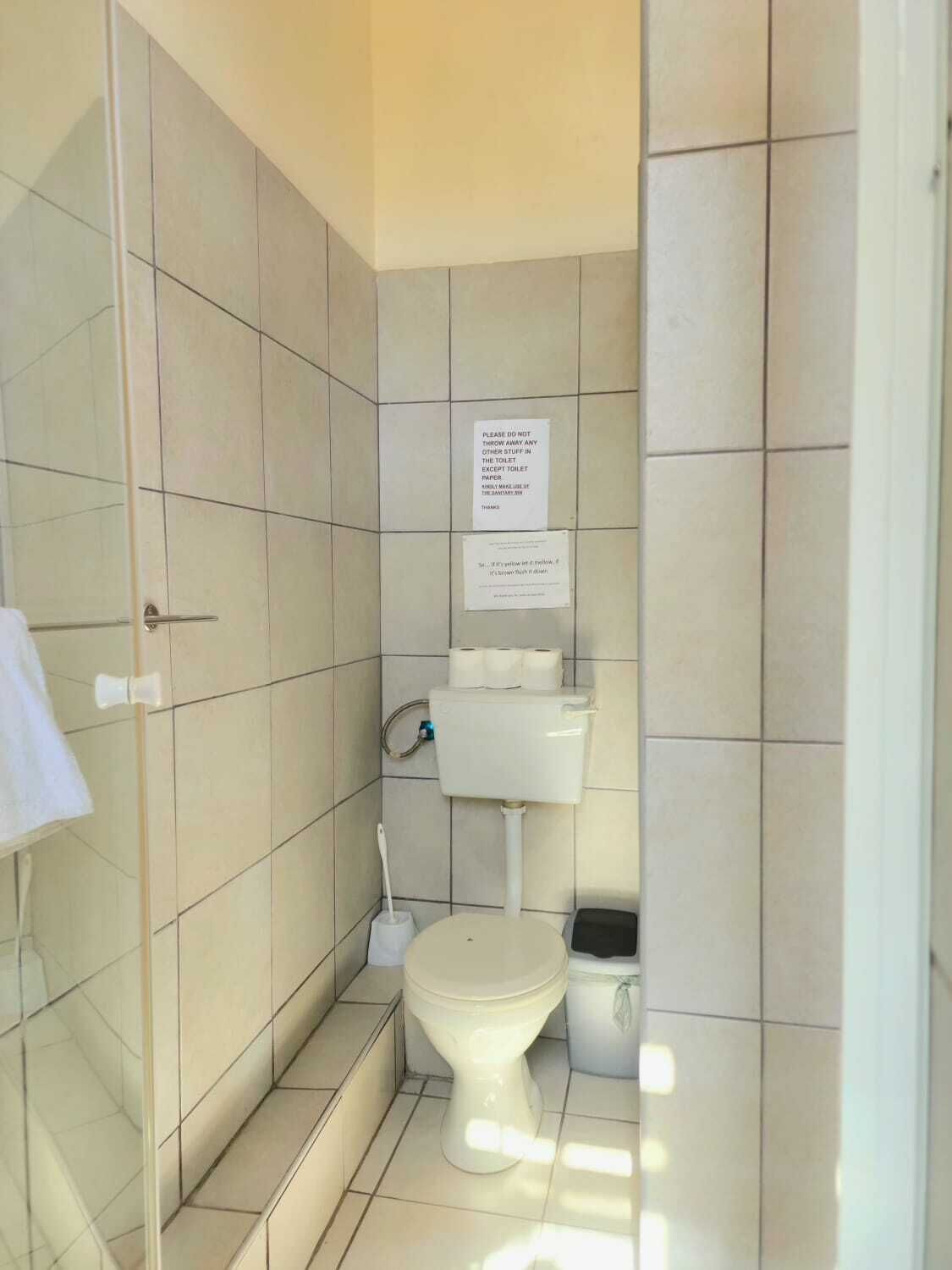 A small, tiled bathroom with a toilet, a roll of toilet paper, and a sign instructing not to throw away any items except toilet paper.