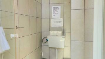 A small, tiled bathroom with a toilet, a roll of toilet paper, and a sign instructing not to throw away any items except toilet paper.