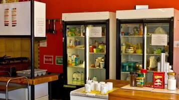 A kitchen area with two refrigerators stocked with food items, a wooden table with kitchenware, and a red wall with a 'Plastic Only' sign.