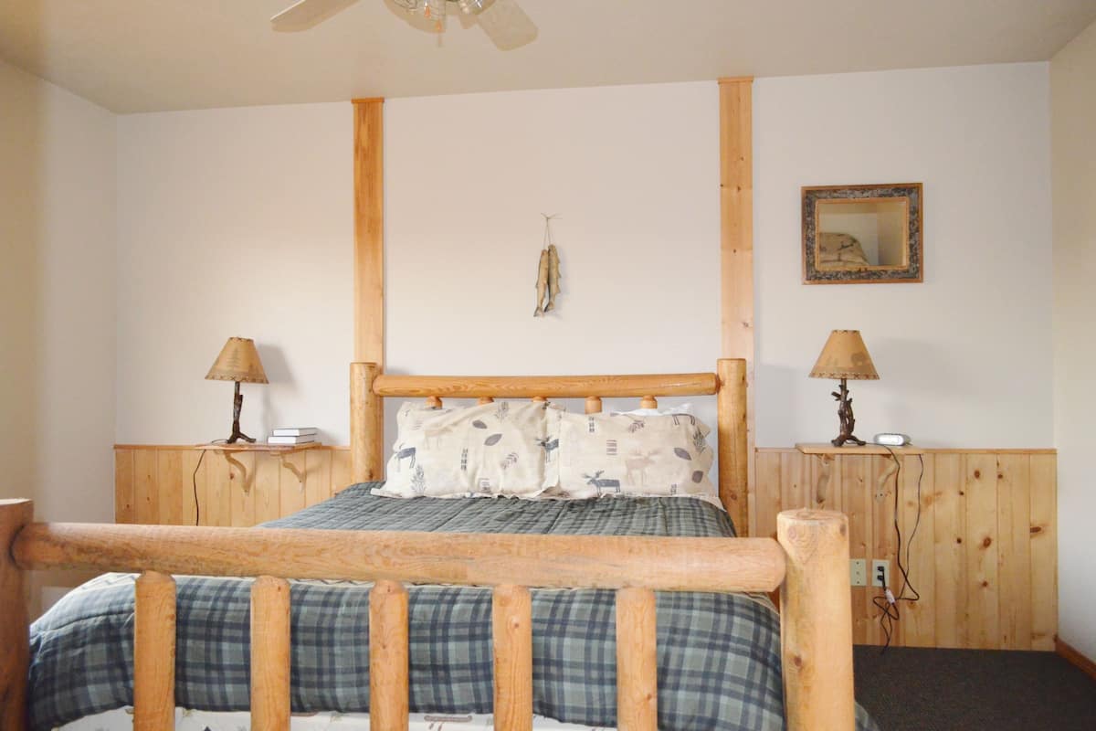 A bedroom with a wooden bed frame, a headboard, two bedside tables with lamps, a wall-mounted mirror, and a hanging ornament.