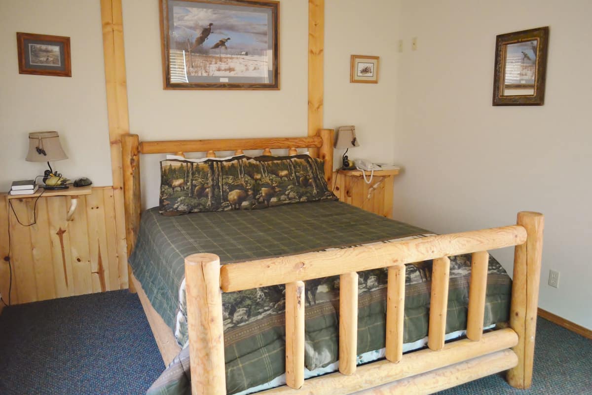 A wooden bed with a green checkered bedspread and matching pillows, a wooden headboard, and a small bedside table with a lamp and a phone.