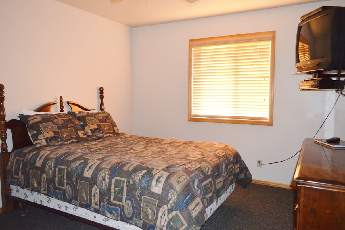 A bedroom with a bed, a wooden headboard, a window with blinds, and a television on a stand.
