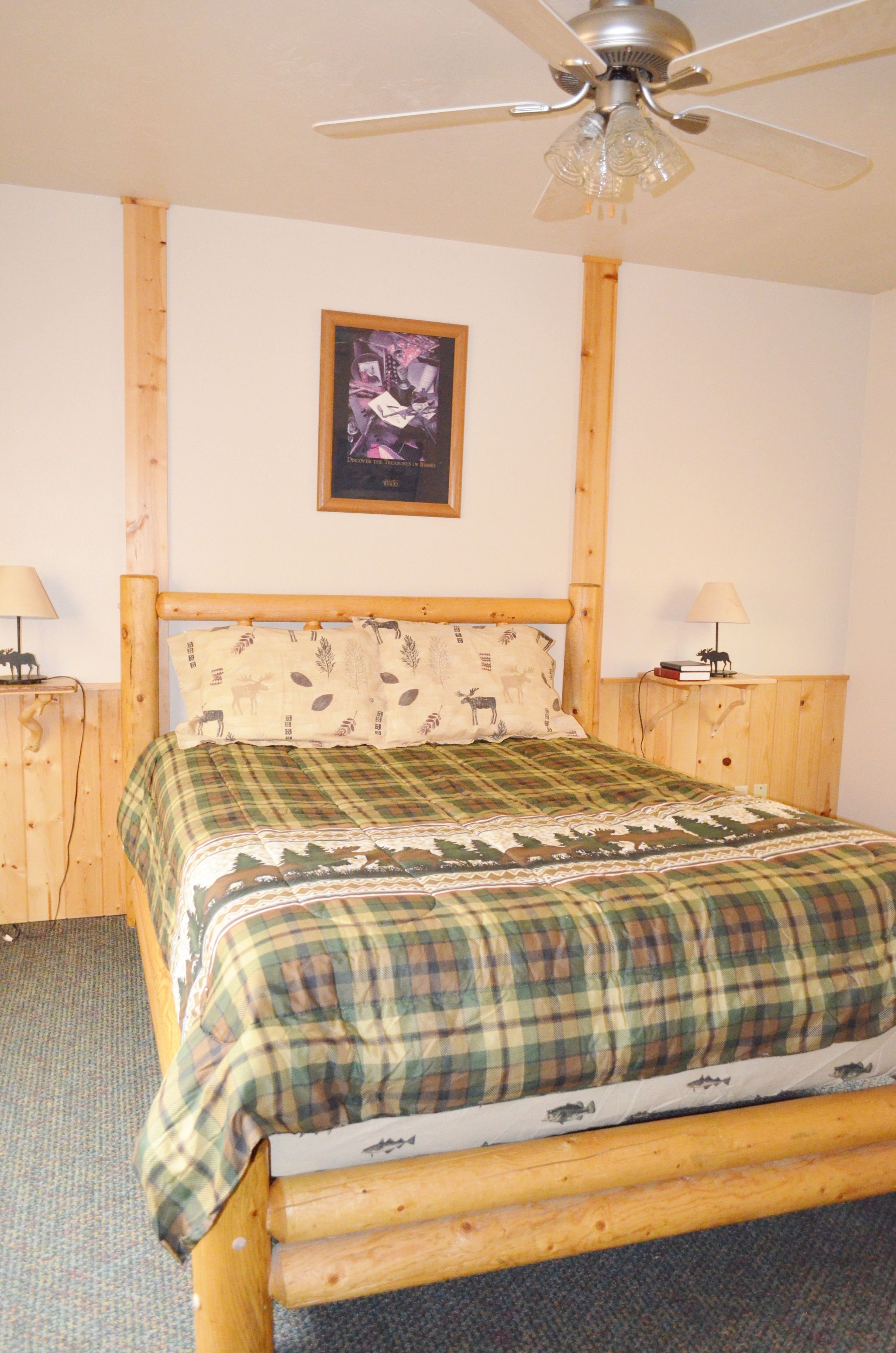 A wooden bed with a plaid bedspread, two bedside tables with lamps, a ceiling fan, and a framed picture on the wall.