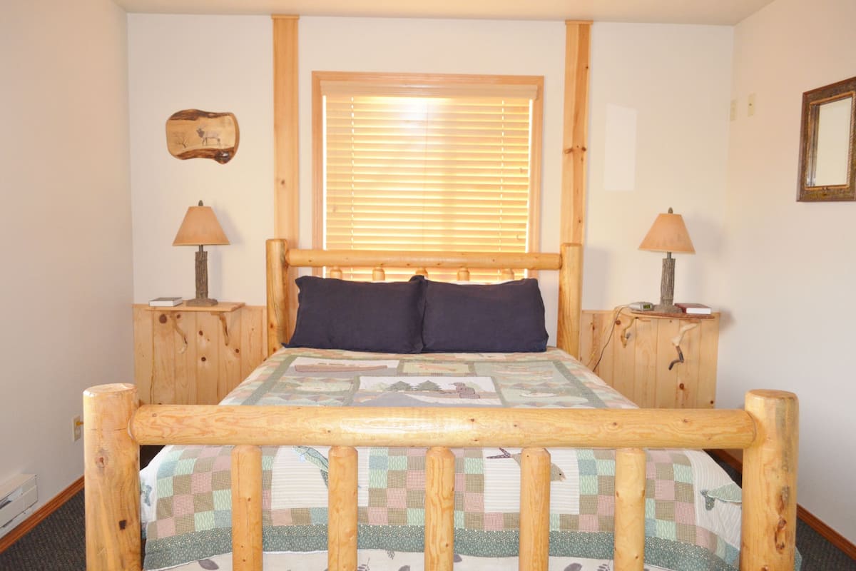 A wooden bed with a quilt, two bedside lamps, a window with blinds, and a wall-mounted picture.