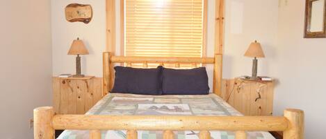 A wooden bed with a quilt, two bedside lamps, a window with blinds, and a wall-mounted picture.